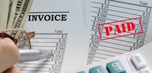 Person holding pen over invoices to the side are a pair of glasses and dollar bills laying on top.