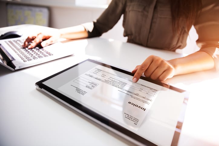 A person reviewing a digital document on a tablet while working with a computer keyboard in the background.