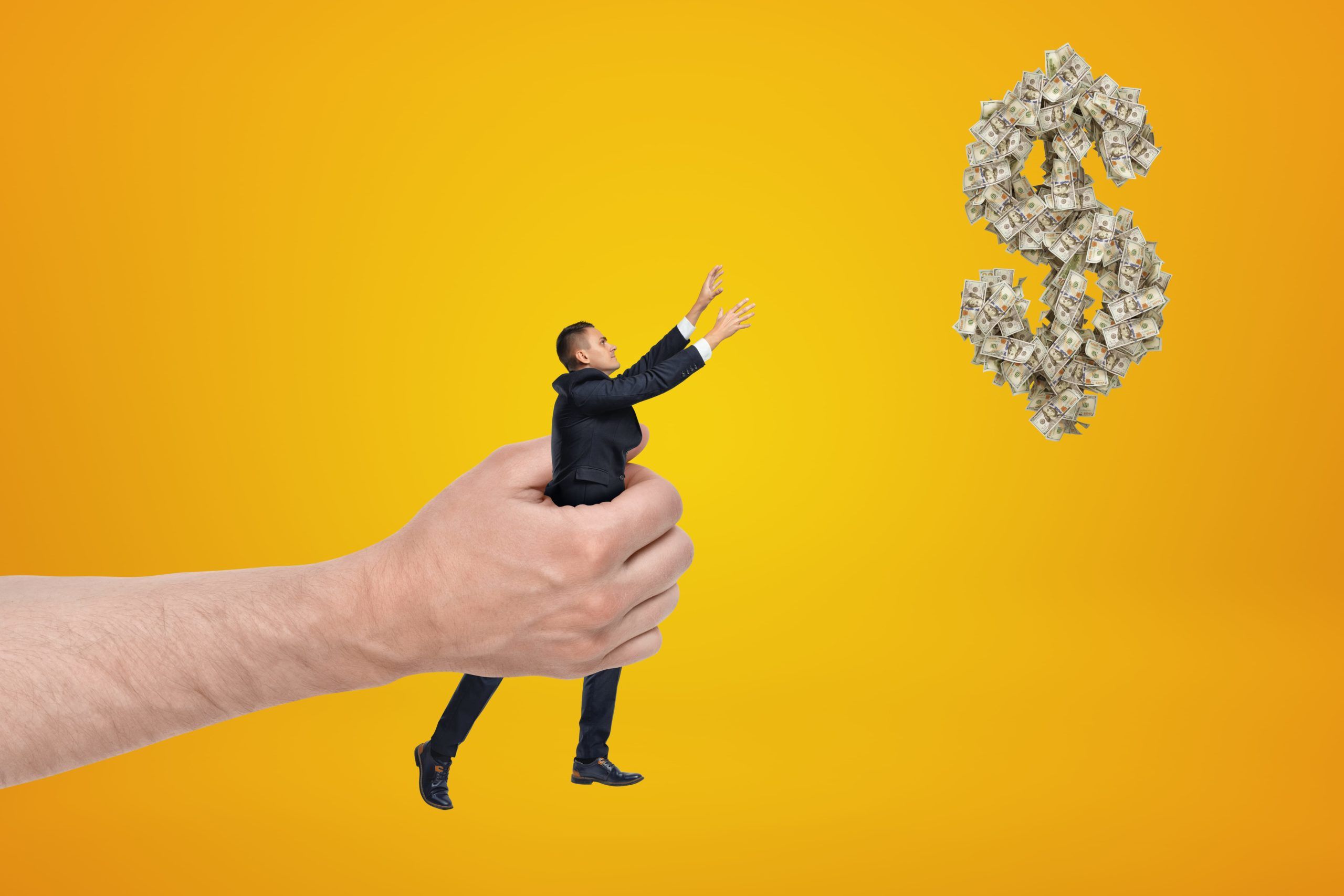 Big hand holding small businessman reaching out with his both hands for dollar symbol made up of dollar bills and floating in air on amber background.