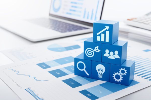 Stacked blue blocks with business icons on a desk, displaying analytics and strategy, with charts and a laptop in the background.