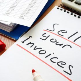 A notebook on a desk with "sell your invoices" written in red marker, alongside a calculator and documents.