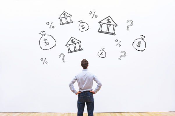 Man contemplating graphics of banks and money bags drawn on a wall, representing financial considerations or decisions.