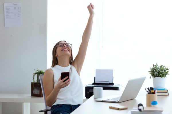 Woman in office raising arm in a gesture of success while holding a smartphone.