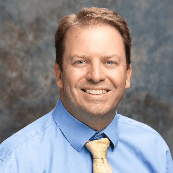 A smiling man with short hair, wearing a blue shirt and a yellow tie against a mottled blue and brown background. This is Lee Nelson.