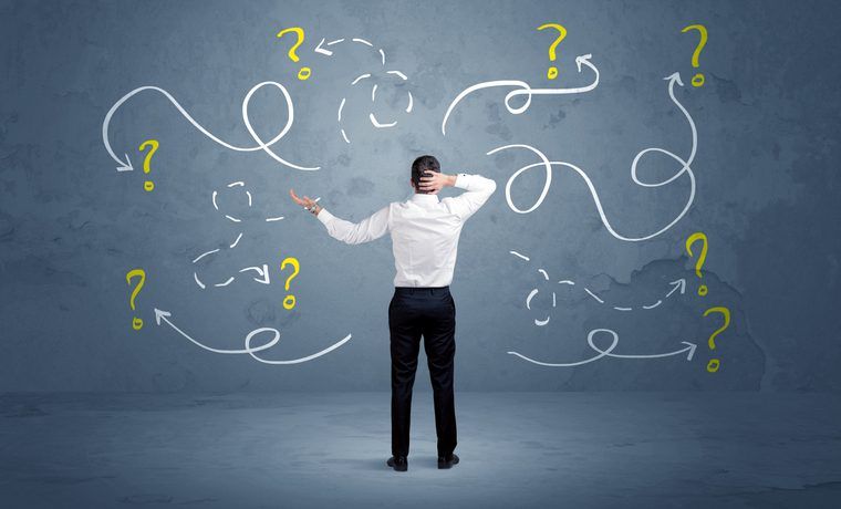 A man in business attire stands with his back to the camera looking at a drawn confusion of question mark doodles on a gray background, suggesting a concept of perplexity or problem-solving in the context of