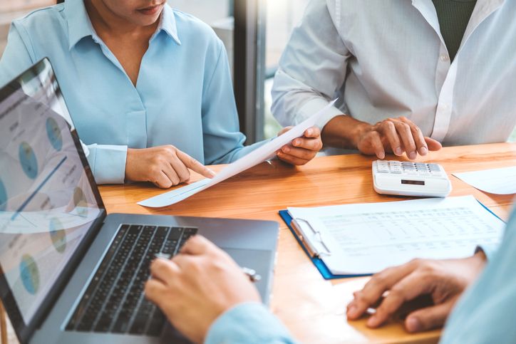 Two professionals analyzing financial documents at a desk with a laptop, printed data sheets, and a calculator to find the right accounts receivable factoring company.