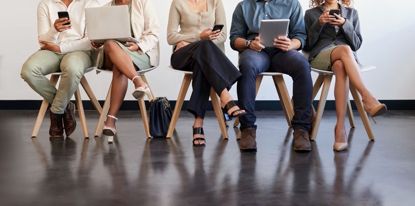 Six individuals seated side by side on chairs, each focused on their own electronic devices to explore the benefits of staffing factoring, with only the lower half of their bodies visible in the frame.