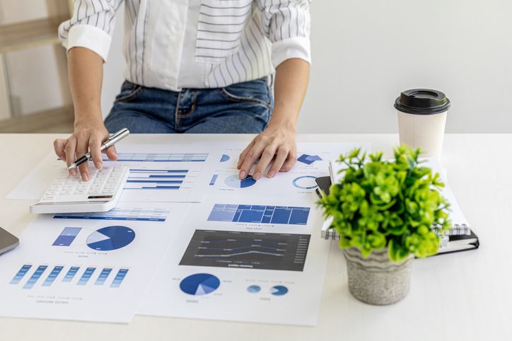 A person sitting at a desk analyzing methods to enhance working capital through financial charts and data with a calculator, pen, coffee cup, and potted plant on the table.