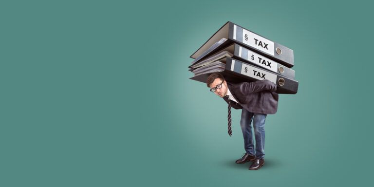 A businessman with a straining expression is carrying a heavy stack of binders showing a tax burden.