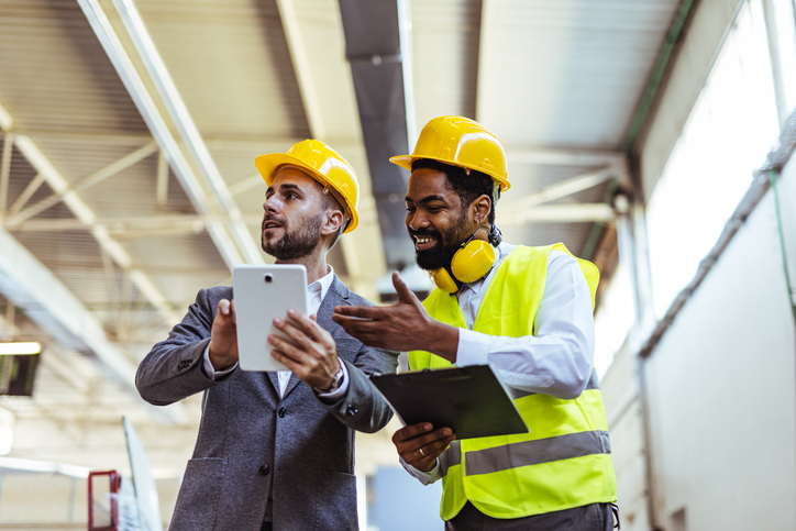 Two manufacturing businessmen collaborating in a warehouse, utilizing a tablet and clipboard to discuss plans accounts receivable factoring to stabilize manufacturing cash flow.